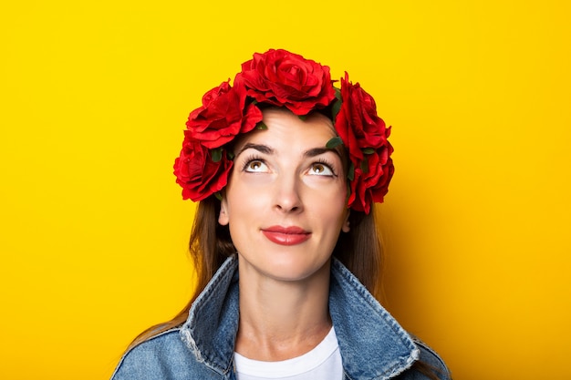 Mujer joven busca con una chaqueta de mezclilla y una corona de flores rojas en la cabeza sobre una pared amarilla.