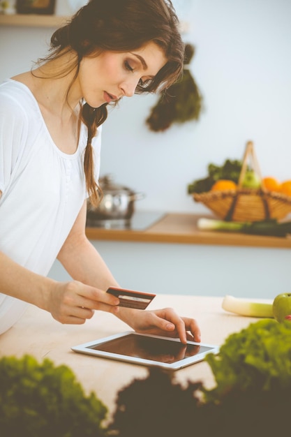 Mujer joven en busca de una nueva receta para cocinar en una cocina. El ama de casa está haciendo compras en línea con una tableta y una tarjeta de crédito.