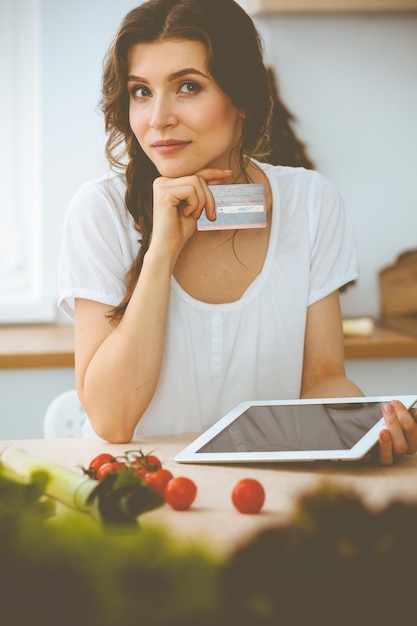 Foto mujer joven buscando una nueva receta para cocinar en una cocina. la ama de casa está haciendo compras en línea por computadora tablet y tarjeta de crédito.