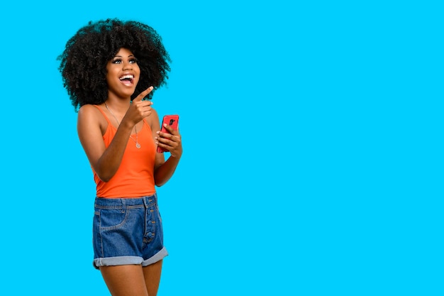 mujer joven con cabello afro sorprendida positivamente apunta a un espacio en blanco en el que sostiene un teléfono inteligente