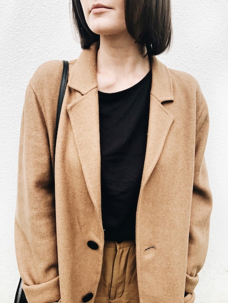 Una mujer joven con cabello castaño, abrigo beige, camisa negra, pantalón marrón de pie contra la pared blanca