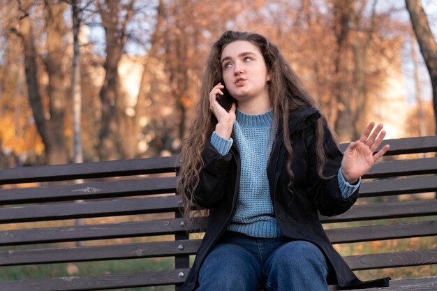 Mujer joven con cabello castaño largo y ondulado se sienta en un banco en el parque e indignada habla por teléfono y gesticula