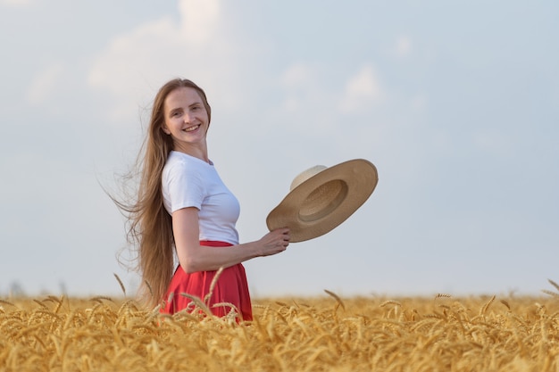 Mujer joven con cabello castaño de pie en el campo de trigo maduro con sombrero. Vacaciones en el campo