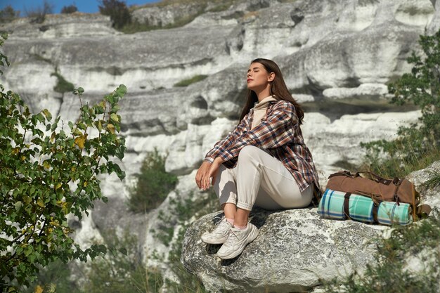 Mujer joven con cabello castaño relajándose sobre una roca, manteniendo los ojos cerrados y disfrutando de su pasatiempo favorito al aire libre.