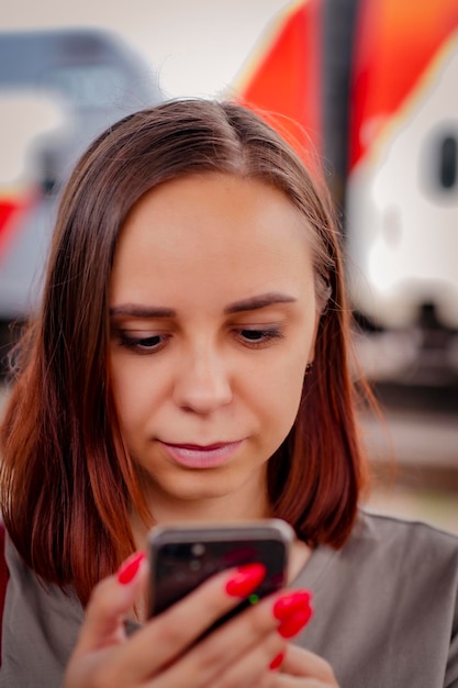 Mujer joven con cabello corto se encuentra en la plataforma navegando por teléfono móvil esperando el tren Retrato de una pasajera usando un teléfono inteligente en el fondo del tren borroso