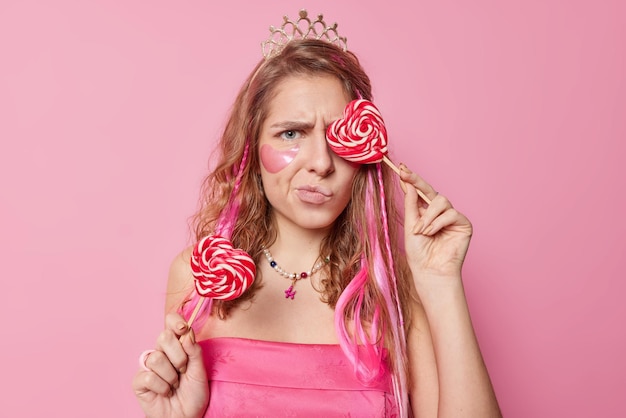 Mujer joven de cabello largo disgustada cubre los ojos con piruleta en forma de corazón frunce el ceño la cara siente descontento aplica parches de hidrogel de belleza debajo de los ojos usa corona y vestido posa contra la pared rosa