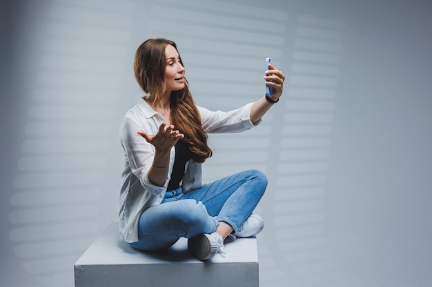 Mujer joven con cabello largo y moreno con una camisa blanca y jeans que tiene una conversación de videollamada por teléfono Una mujer con jeans y una camisa blanca con un teléfono móvil Fondo blanco