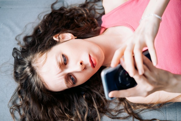 Mujer joven con el cabello largo y rizado se acuesta en el sofá y navega por las redes sociales en su teléfono