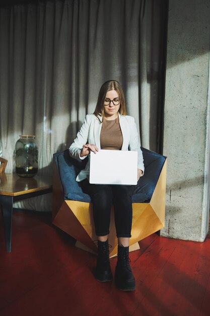 Una mujer joven con cabello largo y rubio con ropa elegante está sentada en una silla y trabajando en una computadora portátil Trabajar en una oficina moderna con ventanas grandes