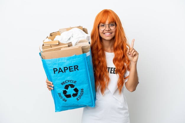 Mujer joven con cabello naranja sosteniendo una bolsa de reciclaje llena de papel para reciclar aislado sobre fondo blanco mostrando y levantando un dedo en señal de lo mejor