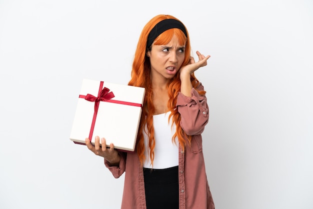 Mujer joven con cabello naranja sosteniendo un regalo aislado sobre fondo blanco frustrado y cubriendo las orejas