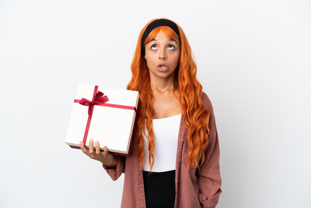 Mujer joven con cabello naranja sosteniendo un regalo aislado sobre fondo blanco mirando hacia arriba y con expresión de sorpresa