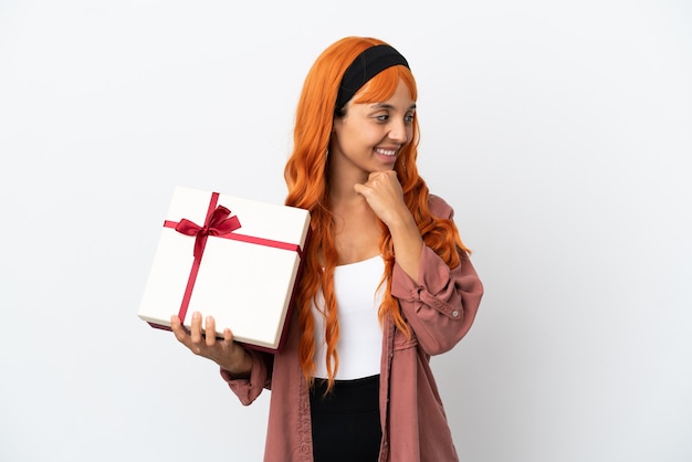 Mujer joven con cabello naranja sosteniendo un regalo aislado sobre fondo blanco mirando hacia el lado y sonriendo