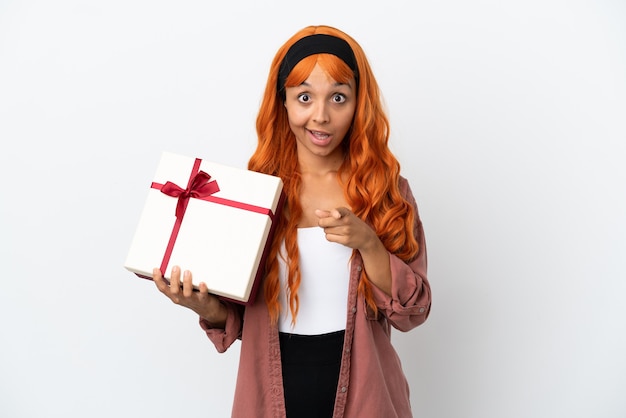 Mujer joven con cabello naranja sosteniendo un regalo aislado sobre fondo blanco sorprendido y apuntando hacia el frente