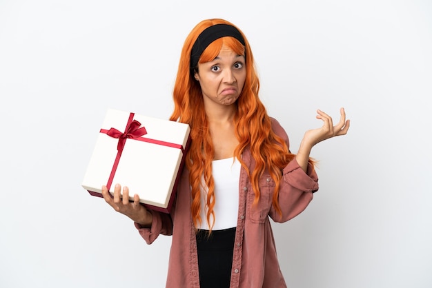 Mujer joven con cabello naranja sosteniendo un regalo aislado sobre fondo blanco teniendo dudas mientras levanta las manos