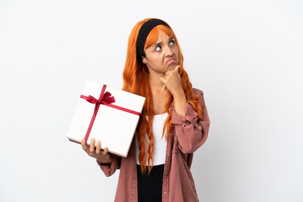 Mujer joven con cabello naranja sosteniendo un regalo aislado sobre fondo blanco teniendo dudas