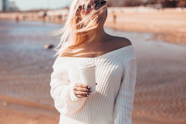 Mujer joven con cabello ondulado rubio sosteniendo una taza de café en la playa