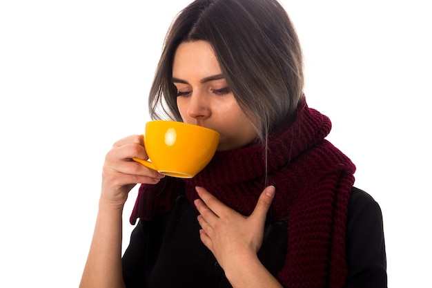 Mujer joven con cabello oscuro en blusa negra con bufanda vinosa bebiendo de una taza amarilla sobre fondo blanco en el estudio