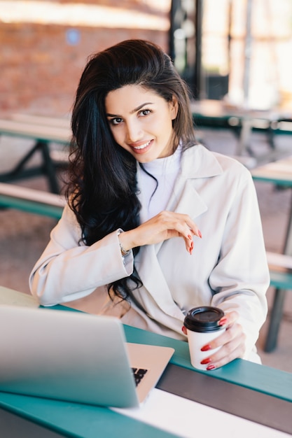 Mujer joven con cabello oscuro con ojos brillantes, labios carnosos y piel sana con bata blanca descansando en el café y navegando por internet