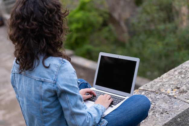 Mujer joven con cabello rizado y anteojos escribiendo en su computadora portátil en el parque en un día nublado