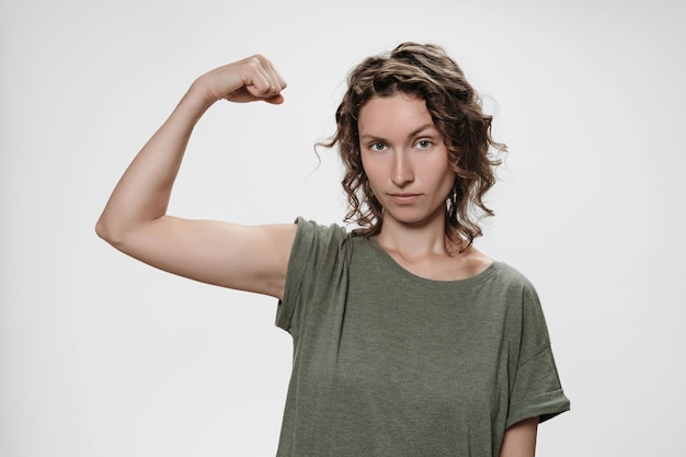 Foto mujer joven de cabello rizado muestra músculo en su mano, se siente orgullosa de ser fuerte y tener fuerza, muestra su gran poder