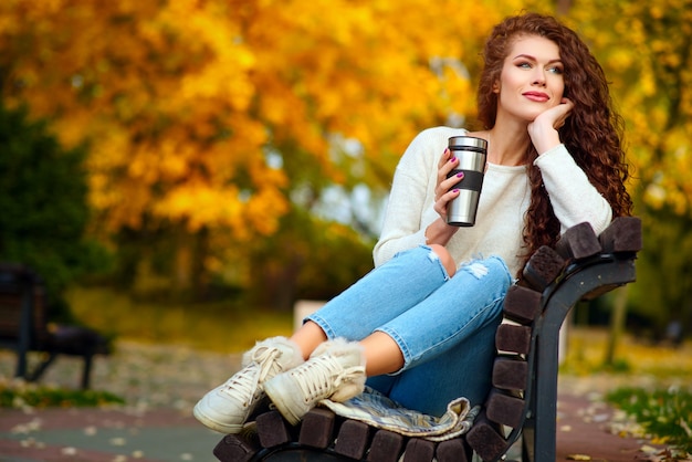 Mujer joven con cabello rizado se sienta en el otoño en un banco en el parque y bebe té de una termocup