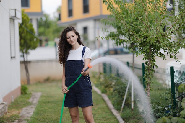 Mujer joven con cabello rizado usando plantas de riego en general en el cuidado del jardín del patio trasero de la casa de campo concepto de vida suburbana de estilo de vida ecológico