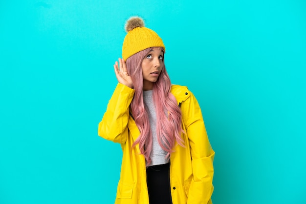 Mujer joven con cabello rosado con un abrigo impermeable aislado sobre fondo azul escuchando algo poniendo la mano en la oreja