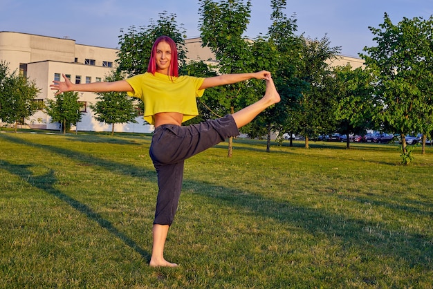 Mujer joven con cabello rosado entrena yoga en un parque de la ciudad en una pose de árbol, vrishasana