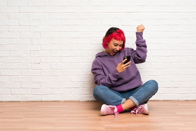 Mujer joven con cabello rosado sentada en el piso con teléfono en posición de victoria