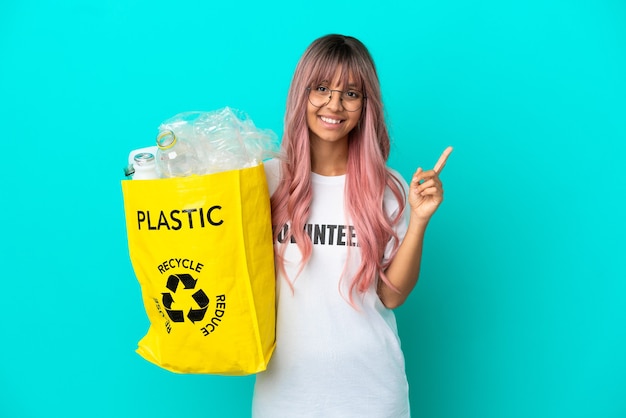 Mujer joven con cabello rosado sosteniendo una bolsa llena de botellas de plástico para reciclar aislado sobre fondo azul apuntando hacia una gran idea