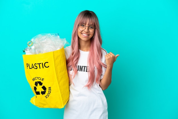 Mujer joven con cabello rosado sosteniendo una bolsa llena de botellas de plástico para reciclar aislado sobre fondo azul apuntando hacia el lado para presentar un producto