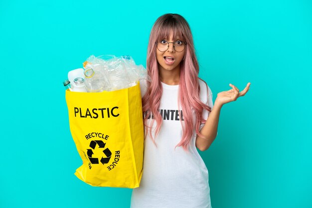 Mujer joven con cabello rosado sosteniendo una bolsa llena de botellas de plástico para reciclar aislado sobre fondo azul con expresión facial conmocionada