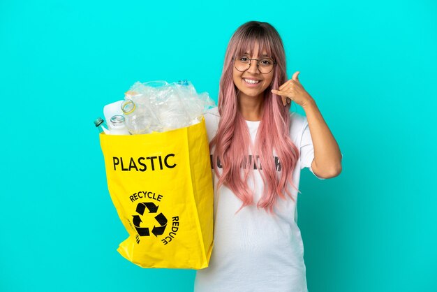 Mujer joven con cabello rosado sosteniendo una bolsa llena de botellas de plástico para reciclar aislado sobre fondo azul haciendo gesto de teléfono. Llámame señal