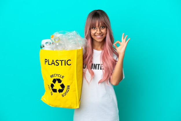 Mujer joven con cabello rosado sosteniendo una bolsa llena de botellas de plástico para reciclar aislado sobre fondo azul mostrando el signo de ok con los dedos