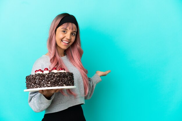 Mujer joven con cabello rosado sosteniendo pastel de cumpleaños aislado sobre fondo azul extendiendo las manos hacia el lado para invitar a venir