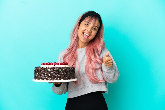 Mujer joven con cabello rosado sosteniendo pastel de cumpleaños aislado sobre fondo azul con los pulgares hacia arriba porque algo bueno ha sucedido