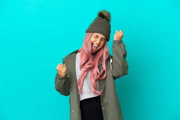 Mujer joven con cabello rosado vistiendo un abrigo impermeable aislado sobre fondo azul celebrando una victoria