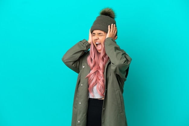 Mujer joven con cabello rosado vistiendo un abrigo impermeable aislado sobre fondo azul estresado abrumado