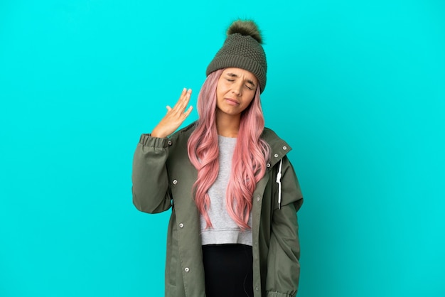 Mujer joven con cabello rosado vistiendo un abrigo impermeable aislado sobre fondo azul con expresión cansada y enferma