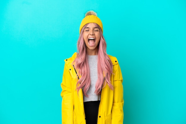 Mujer joven con cabello rosado vistiendo un abrigo impermeable aislado sobre fondo azul gritando al frente con la boca abierta