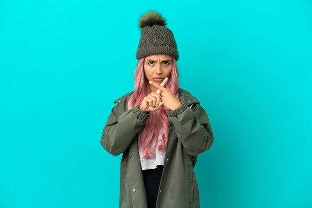 Mujer joven con cabello rosado vistiendo un abrigo impermeable aislado sobre fondo azul haciendo gesto de parada con la mano para detener un acto