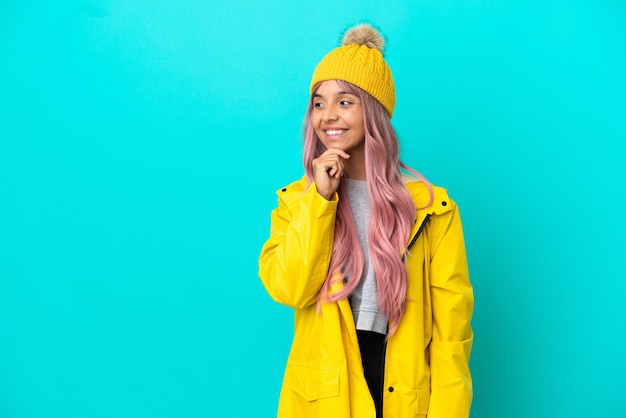 Mujer joven con cabello rosado vistiendo un abrigo impermeable aislado sobre fondo azul mirando hacia el lado y sonriendo