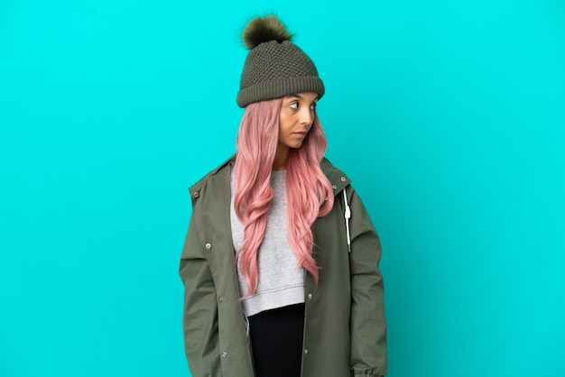 Mujer joven con cabello rosado vistiendo un abrigo impermeable aislado sobre fondo azul mirando hacia el lado