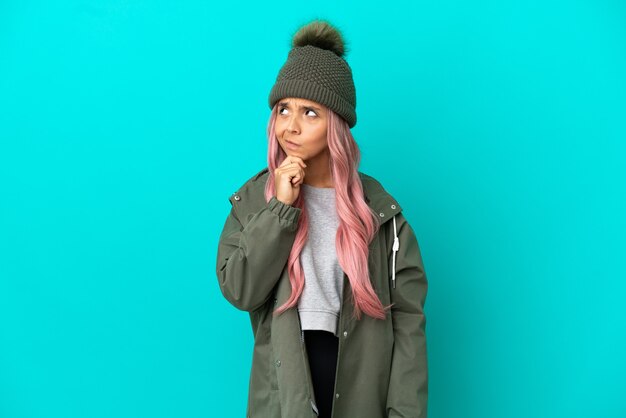 Mujer joven con cabello rosado vistiendo un abrigo impermeable aislado sobre fondo azul teniendo dudas y pensando