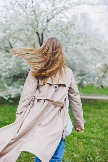 Una mujer joven con cabello rubio disfruta de un floreciente jardín primaveral Vacaciones de primavera de viaje Estilo de moda Una mujer con gafas de sol y una gabardina beige corre por un parque florido Enfoque de selección