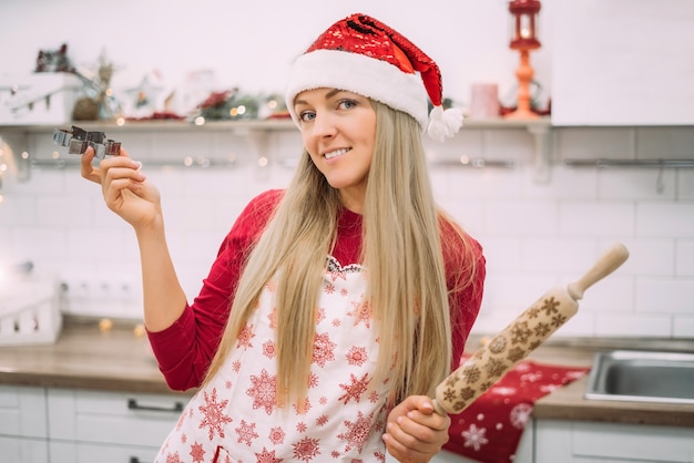 Una mujer joven con cabello rubio se encuentra en la cocina vistiendo un delantal y una bandeja de galletas de jengibre en un gorro de Papá Noel