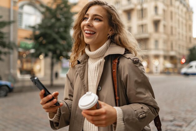 Mujer joven con cabello rubio rizado usando el teléfono con una taza de café en las manos en las calles de la ciudad