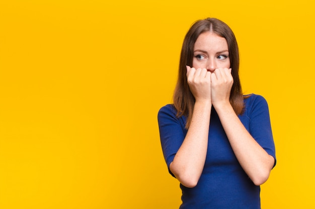 Mujer joven de cabeza roja que parece preocupada, ansiosa, estresada y asustada, mordiéndose las uñas y mirando hacia el espacio de copia lateral contra la pared plana