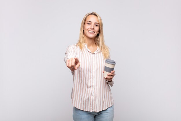 Mujer joven con un café apuntando a la cámara con una sonrisa satisfecha, confiada y amistosa, eligiéndote